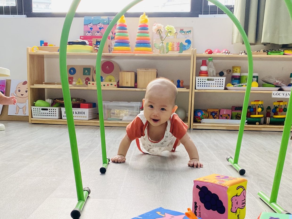 Bé lớp Baby bò qua vòm xanh trong phòng học, phía sau là kệ đồ chơi gỗ