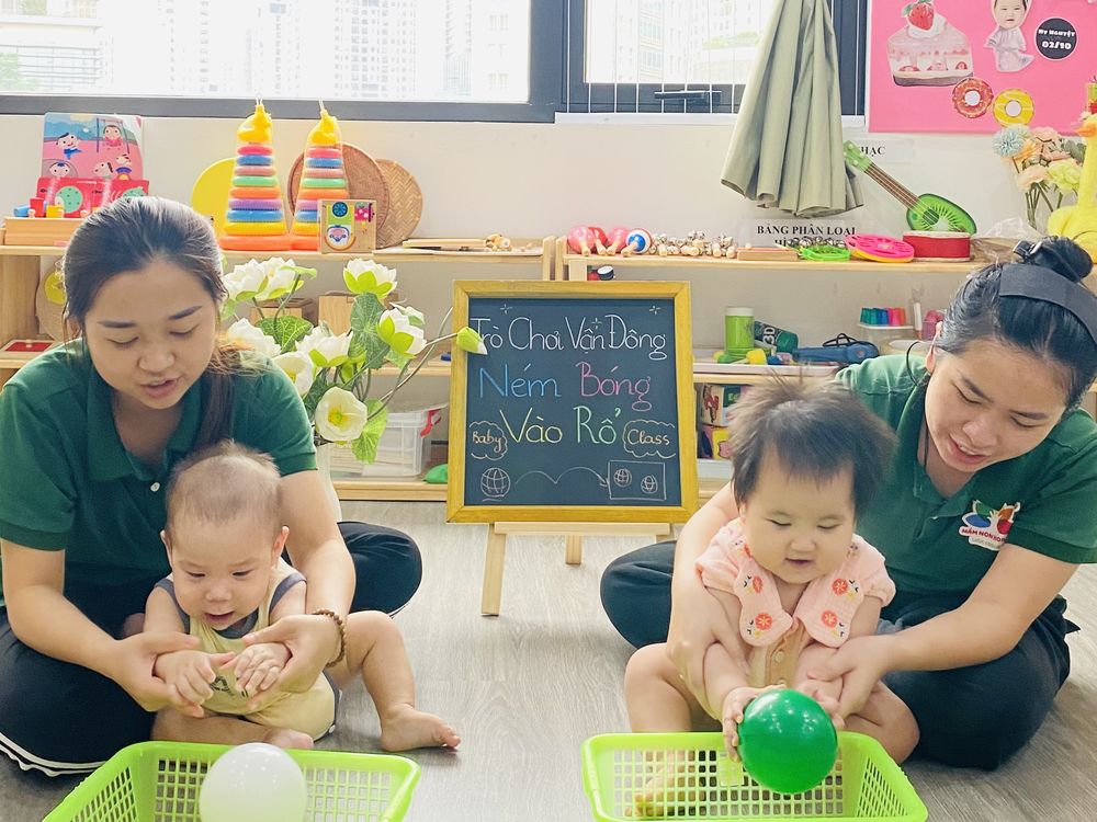 Hai cô giáo ngồi cùng hai bé lớp Baby chơi ném bóng vào rổ, bảng lớp ghi Trò chơi vận động - Ném bóng vào rổ - Baby Class
