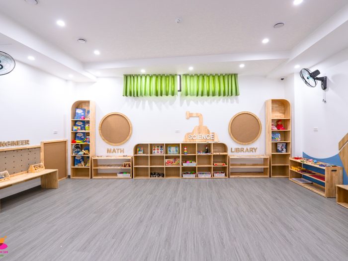Phòng học của trường với kệ gỗ, rèm xanh và sàn rộng cho bé vận động
