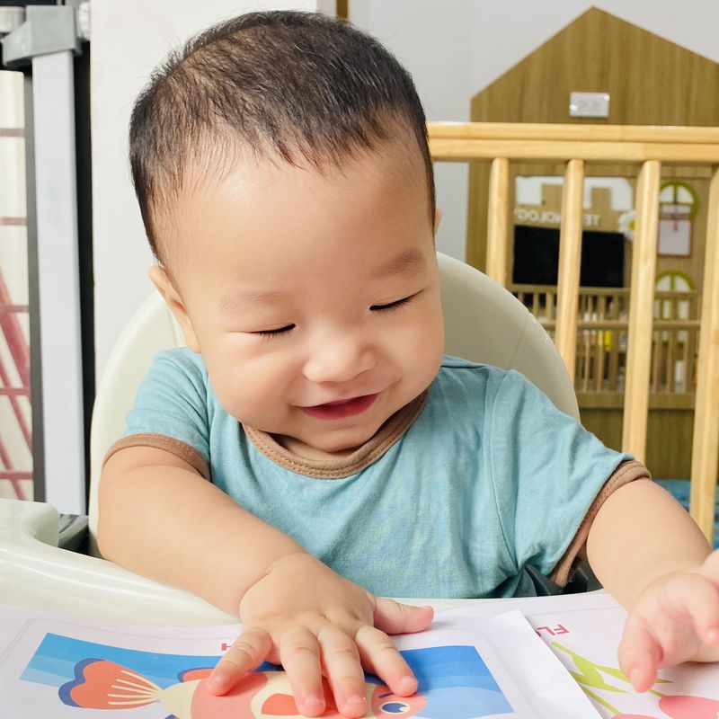 Bé lớp Baby ngồi ghế ăn, mỉm cười, hai tay đặt lên tấm thẻ hình con cá