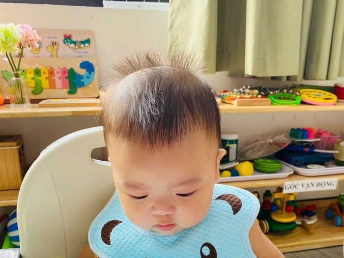 Bé lớp Baby đeo yếm xanh ngồi ghế ăn, phía sau là kệ đồ chơi