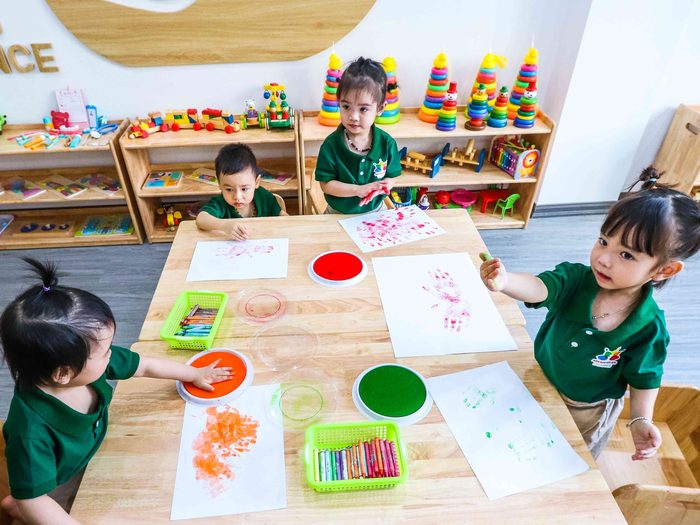 Nhóm bé mặc đồng phục xanh ngồi quanh bàn, in dấu tay bằng màu lên giấy trong lớp học